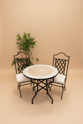 A 3-piece outdoor bistro set featuring a round mosaic table with a black metal frame and two matching black metal chairs with white cushions, set against a plain white wall with a potted plant.