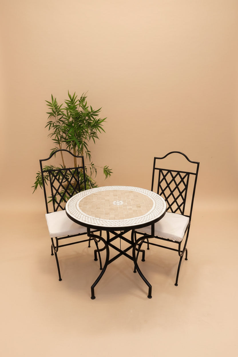 A 3-piece outdoor bistro set featuring a round mosaic table with a black metal frame and two matching black metal chairs with white cushions, set against a plain white wall with a potted plant.